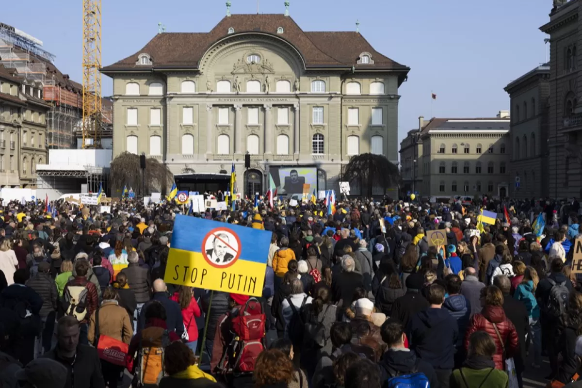 Președintele ucrainean Volodymyr Zelensky este afișat pe un ecran în timpul unei demonstrații împotriva invaziei ruse a Ucrainei în fața Băncii Naționale Elvețiene, în spate, și a clădirii parlamentului elvețian, în dreapta, la Berna, Elveția, 19 martie 2022.